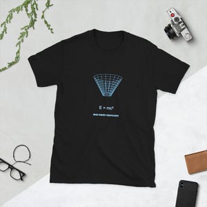 Puede incluir: Una camiseta negra de manga corta con un gráfico de alambre azul claro de un cono de pozo gravitatorio, la ecuación E = mc² en texto blanco y las palabras MASS-ENERGY EQUIVALENCE impresas en azul claro en el centro del pecho.