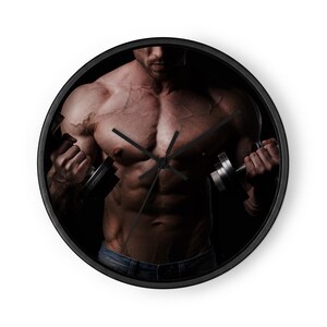 May include: A round wall clock with a black frame featuring a photographic print of a muscular torso holding two metal dumbbells. The clock face has three thin black hands positioned against the dark, high-contrast image of the upper body.