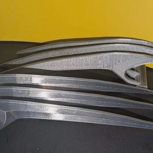 Può includere: Un set di due oggetti di scena a forma di artiglio a tre punte di colore argento. Ogni oggetto presenta tre lame lunghe, curve e appuntite collegate a una base. La superficie mostra una finitura stratificata tipica della stampa 3D.