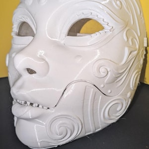 May include: A white, full-face mask featuring intricate, raised swirl and scroll patterns across the forehead and cheeks. The mask has almond-shaped eye openings, a defined nose, and a mouth area with visible teeth, all moulded in a smooth finish.
