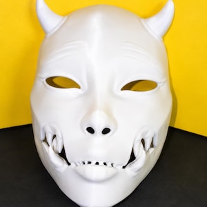 Könnte beinhalten: Eine einfarbig weiße Maske mit zwei markanten Hörnern an der Oberseite, mandelförmigen Augenöffnungen und einem detaillierten Mundbereich mit sichtbaren Fangzähnen und Zähnen. Die Maske hat eine glatte, matte Oberfläche und ein spitzes Kinn.