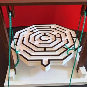 May include: A white octagonal maze game suspended by green strings from a brown rectangular frame. The maze features black pathways and circular holes on a white base. The structure is mounted on a white platform, with a small coin visible nearby.