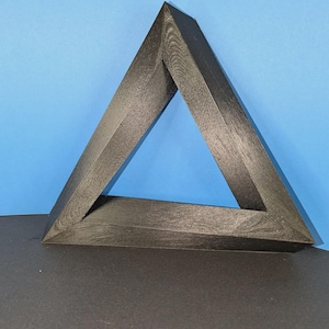 May include: A black, three-dimensional triangular object with a textured surface. The structure features a hollow centre and sharp, angular edges, creating a geometric form that appears to be a solid, continuous loop with a matte finish.