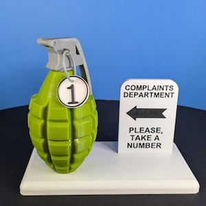 May include: A lime green hand grenade replica with a grey top lever and a circular white tag featuring the black number 1. Next to it stands a white sign with black text reading COMPLAINTS DEPARTMENT, a left-pointing arrow, and PLEASE, TAKE A NUMBER.