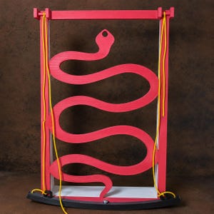 May include: A rectangular red frame featuring a cutout of a snake shape in the centre. Two bright yellow cords hang vertically from the top corners, connecting to a black curved base at the bottom. The structure has a white interior edge.