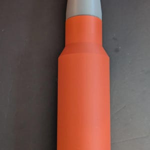 Könnte beinhalten: Ein zylindrisches Objekt in Form eines großkalibrigen Geschosses mit einer matten orangefarbenen Hülse und einer glatten, spitz zulaufenden hellgrauen Projektilspitze. Der Gegenstand hat einen ausgeprägten Hals und einen flachen Boden.