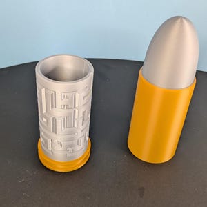 May include: A silver cylindrical container featuring a recessed maze pattern on its exterior, paired with a separate yellow casing topped with a silver bullet-shaped cap. The two components are displayed side-by-side as a modular storage unit.