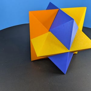 May include: A geometric sculpture composed of interlocking polyhedral shapes in vibrant orange, yellow, and blue. The object features multiple triangular faces meeting at sharp vertices, creating a complex three-dimensional star-like structure.