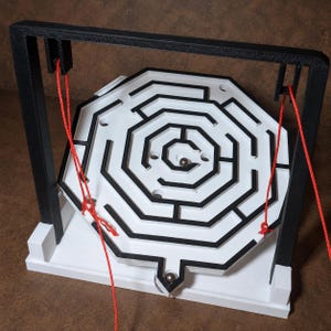 May include: A white octagonal maze game with black walls suspended by red strings from a black frame. The game features a central metal ball bearing and circular holes throughout the path. The entire structure is mounted on a white base.
