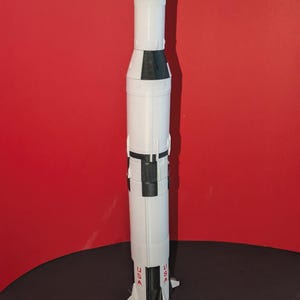 May include: A vertical model of a Saturn V rocket featuring a white cylindrical body with black segmented bands. The lower section displays the red text USA on two sides. The base includes multiple engine nozzles and small stabilizing fins.