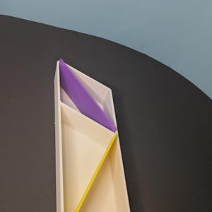 Puede incluir: Una bandeja blanca alargada de múltiples compartimentos con una forma geométrica angular. La bandeja cuenta con un divisor interno de color morado brillante y otro divisor interno de color amarillo vibrante, creando tres secciones triangulares distintas.