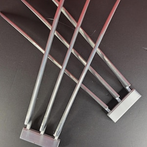 Könnte beinhalten: Zwei Sets aus je drei silberfarbenen, spitz zulaufenden, klingenartigen Elementen, die an rechteckigen Basen befestigt sind. Jedes Set besteht aus drei langen, schlanken, metallisch wirkenden Zinken, die aus einem flachen Griffstück ragen.
