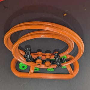 May include: An orange plastic stand featuring two large circular rings connected to a base. The structure includes several small black and orange spools arranged in a row, with bright green circular accents positioned along the base frame.