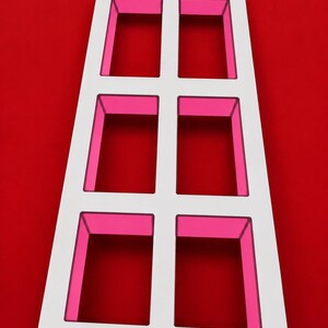 Puede incluir: Un marco rectangular blanco con seis compartimentos rectangulares abiertos con paredes interiores de color rosa vibrante. La estructura está orientada verticalmente e incluye dos pequeños orificios circulares en las esquinas superiores.