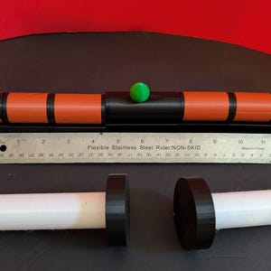 Op de afbeelding: Een cilindrisch oranje en zwart gereedschap met een kleine groene bol aan de bovenkant, geplaatst boven een roestvrijstalen liniaal van 30 cm. Onder de liniaal bevinden zich twee witte cilindrische onderdelen, elk met een zwarte ronde eindkap.