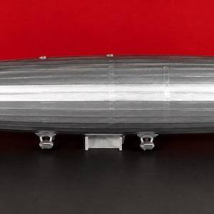 May include: A silver-colored model airship with a long, cylindrical, tapered body and a pointed nose. The airship features horizontal and vertical tail fins at the rear, small undercarriage structures along the bottom, and rests on a white stand.
