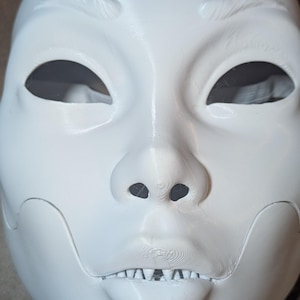 以下が含まれることがあります： 滑らかで様式化された人間のような外観を持つ、白い3Dプリントのフルフェイスマスク。アーモンド型の目の切り抜き、はっきりとした鼻、小さな歯が見える分離された顎のラインが特徴です。表面にはわずかな水平のプリントラインが見えます。