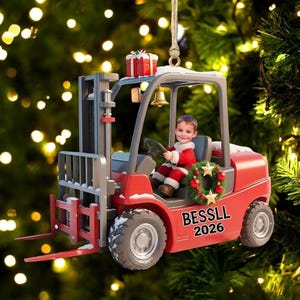 May include: A red forklift Christmas ornament featuring a child in a Santa suit sitting in the driver seat. The forklift has a small wreath on the side, a gift box on top, and the text BESSLL 2026 printed in black on the side of the vehicle body.