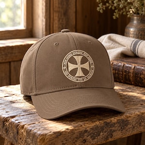 Peut inclure: Une casquette de baseball beige avec un emblème circulaire brodé sur le devant. L'emblème présente une croix au centre entourée du texte latin "NON NOBIS DOMINE NON NOBIS SED NOMINI TUO DA GLORIAM" inscrit dans une bordure circulaire.