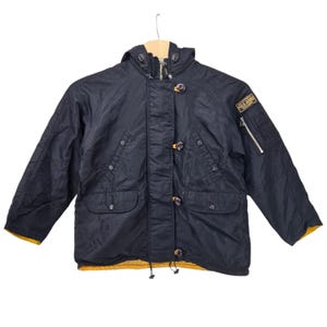 May include: A navy blue hooded jacket featuring a front zipper closure with a buttoned storm flap, two lower flap pockets, and a zippered utility pocket on the left sleeve. A rectangular patch on the chest reads Polo Jeans Co. Ralph Lauren.