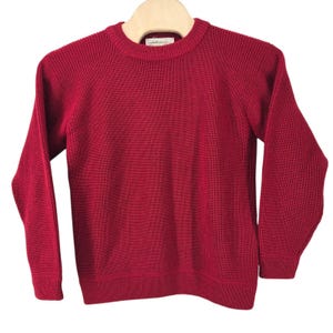 May include: A solid deep red long-sleeve jumper featuring a classic crew neckline and a ribbed knit texture. The garment is displayed on a wooden hanger and includes a small white interior brand label at the centre of the neckline collar.