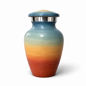 May include: A cremation urn featuring a smooth, gradient finish that transitions from a deep red at the base through orange and yellow to a light blue at the top. The vessel has a rounded body and a matching lid with a silver metal band.