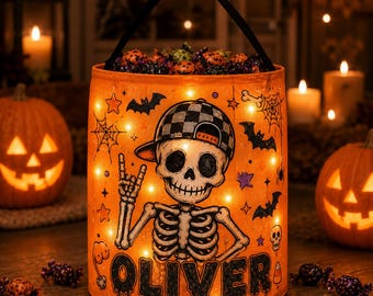 Puede incluir: Un cubo de Halloween cilíndrico de color naranja con asa negra, que muestra un esqueleto con gorra de béisbol a cuadros haciendo un gesto con la mano. El cubo está iluminado con pequeñas luces y decorado con murciélagos, telarañas y el nombre OLIVER.