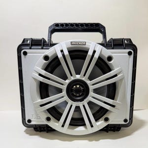 Puede incluir: Un maletín rígido negro portátil con un altavoz circular Kicker grande montado en la parte frontal. El altavoz tiene una rejilla blanca de múltiples radios y un cono central negro. Un asa negra está fijada en la parte superior.