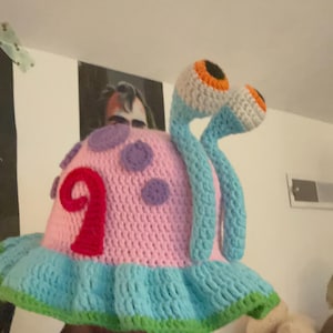 Könnte beinhalten: Ein gehäkelter Fischerhut in Form einer Schnecke mit einem rosafarbenen Gehäuse, das violette Punkte und ein rotes Spiralmuster aufweist. Der Hut hat eine hellblaue, gerüschte Krempe mit grünem Rand sowie zwei hellblaue Stiele mit weiß-orangen Augen.