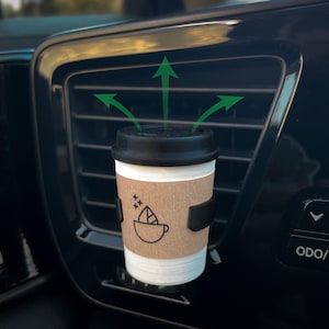 May include: A miniature coffee cup air freshener attached to a black car air vent. The cup has a white base, a tan cardboard-style sleeve with a black line drawing of a coffee cup and leaf, and a black lid. Three green arrows point upward.