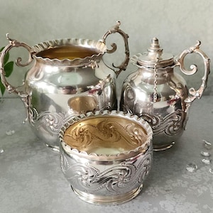May include: A three-piece silver-plated tea set consisting of a large sugar bowl, a smaller open bowl, and a lidded creamer. Each piece features ornate embossed scrollwork, scalloped rims, and decorative handles with a polished metallic finish.