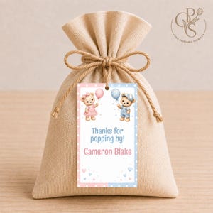 Op de afbeelding: Een beige stoffen zakje met een jute trekkoord en een rechthoekig cadeaulabel. Het label toont twee teddyberen met ballonnen, met de tekst "Thanks for popping by!" en de naam "Cameron Blake" gedrukt in blauw en roze.