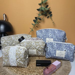 May include: A collection of four quilted cosmetic pouches in olive green and blue floral patterns, each featuring a white label with the text Nuvika. Two pouches have white lace trim, and the set includes a black lipstick and a pink lip gloss.