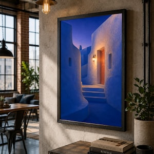 May include: A rectangular framed wall art print featuring a narrow, winding alleyway between white buildings at twilight. The scene is bathed in deep blue shadows, contrasted by a warm, glowing wall-mounted lantern illuminating a wooden door.