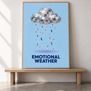May include: A framed wall art print featuring a light blue background with a central disco ball cloud raining down colorful rectangular confetti pieces. Below the cloud, the text EMOTIONAL WEATHER is printed in bold, dark blue capital letters.