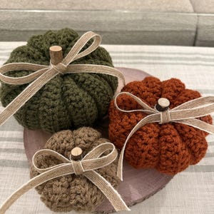 May include: Three crocheted pumpkins in olive green, burnt orange, and tan. Each pumpkin features a small wooden stem and a decorative beige ribbon tied in a bow. The pumpkins are arranged together on a circular wooden slice base.