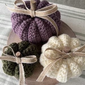 May include: Three crocheted pumpkins in purple, olive green, and cream colours. Each pumpkin features a small wooden stem and a decorative tan ribbon tied in a bow around the top. The pumpkins are arranged together on a round wooden base.