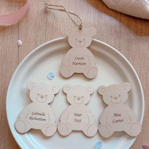 May include: Four light-colored wooden bear-shaped ornaments with engraved text. One ornament has a jute twine loop. The engravings read Lucas Harrison, Gabriella Richardson, Your Text, and Mia Carter. The bear shapes feature simple etched facial details.