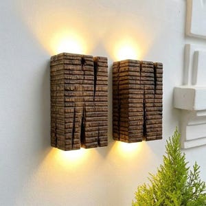 Puede incluir: Dos apliques de pared rectangulares fabricados con bloques de madera de color marrón oscuro y textura rugosa. Cada lámpara presenta una hendidura vertical en el centro y emite una luz cálida desde los bordes superior e inferior.
