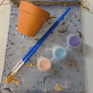 May include: A terracotta plant pot, two blue paintbrushes, and a strip of three small paint pots containing peach, light blue, and purple paint, all resting on a sheer blue drawstring bag decorated with gold glitter moons and stars.