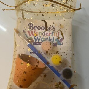 May include: A craft kit in a gold star-patterned organza drawstring bag containing a small terracotta pot, a blue paintbrush, and three small paint pots in peach, yellow, and black. A card inside reads Brooke's Wonderful World and Tiny Crafts.