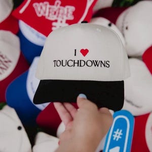 Könnte beinhalten: Eine weiße Baseballkappe mit schwarzem Schirm, die den gestickten Text I gefolgt von einem roten Herzsymbol und dem Wort TOUCHDOWNS in schwarzer Schrift zeigt. Die Kappe hat eine strukturierte Krone und einen Knopf oben, vor einem Hintergrund aus Sportartikeln.