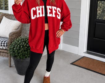 Könnte beinhalten: Ein leuchtend rotes Sweatshirt mit Reißverschluss und dem Wort CHIEFS in großen weißen Blockbuchstaben auf der Brust. Das Kleidungsstück hat lange Ärmel, einen Rundhalsausschnitt und eine lockere Passform, getragen über einem schwarzen Oberteil und einer schwarzen Leggings mit braunen Schuhen.