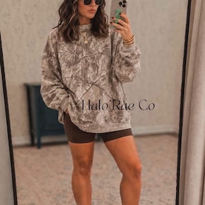 May include: A brown and white trucker hat with the text 'Halo Rae Co', a grey and white camouflage print sweatshirt, dark brown biker shorts, white crew socks, and beige athletic sneakers. The outfit includes round sunglasses and a gold star necklace.