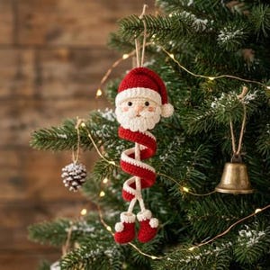 Può includere: Un ornamento di Babbo Natale all'uncinetto con un cappello rosso e bianco, una barba bianca e un lungo corpo a spirale rosso e bianco che termina con due piccoli stivali rossi. La figura è appesa a un filo sottile su un ramo di pino.