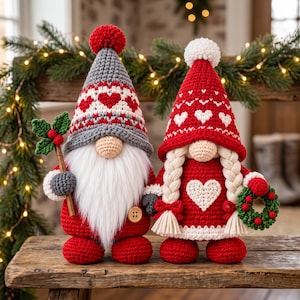 Puede incluir: Dos gnomos de ganchillo con diseños festivos. El gnomo de la izquierda tiene un gorro gris con corazones rojos, una larga barba blanca y sostiene una rama de acebo. El de la derecha tiene un gorro rojo con corazones blancos, trenzas y una corona.