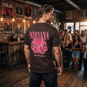 May include: A dark brown short-sleeve t-shirt featuring a pink graphic on the back. The graphic displays the text NIRVANA in large letters above a stylised pink heart shape and the text Heart-Shaped Box in a cursive font at the bottom.