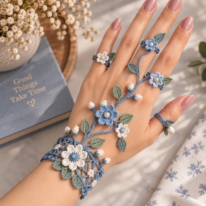 Op de afbeelding: Een blauw en wit gehaakte handbloemarmband met ingewikkelde bloemmotieven, groene blaadjes en kleine parelachtige kraaltjes. Het ontwerp verbindt een brede polsband met meerdere vingerringen door middel van delicaat haakwerk.