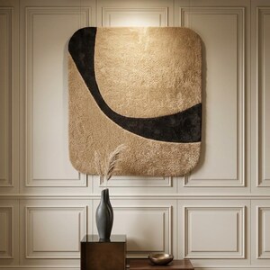 May include: A large, square, textured wall hanging with rounded corners, featuring a beige base with a sweeping black curved design. Below, a dark gray vase with dried grass sits on a dark metal cube table next to a low wooden block.