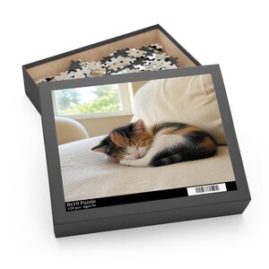 May include: A black puzzle box containing loose pieces, featuring a printed image of a sleeping calico kitten on a beige surface. The box lid displays the text 8x10 Puzzle 120 pcs Ages 9+ and a barcode. The interior of the box holds scattered puzzle pieces.